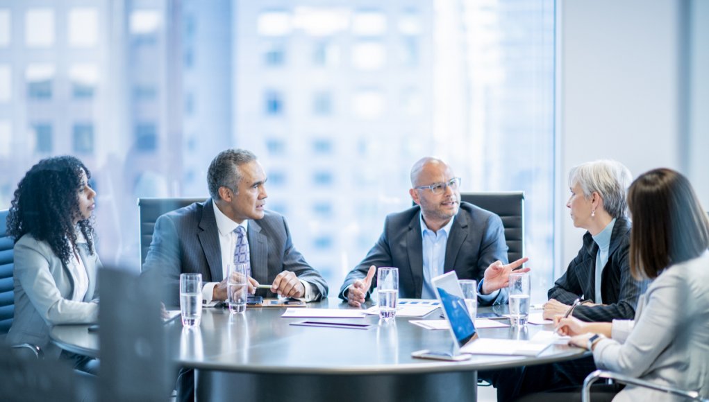 A group of five businesspeople are sitting around an office table. They are talking while having a meeting.