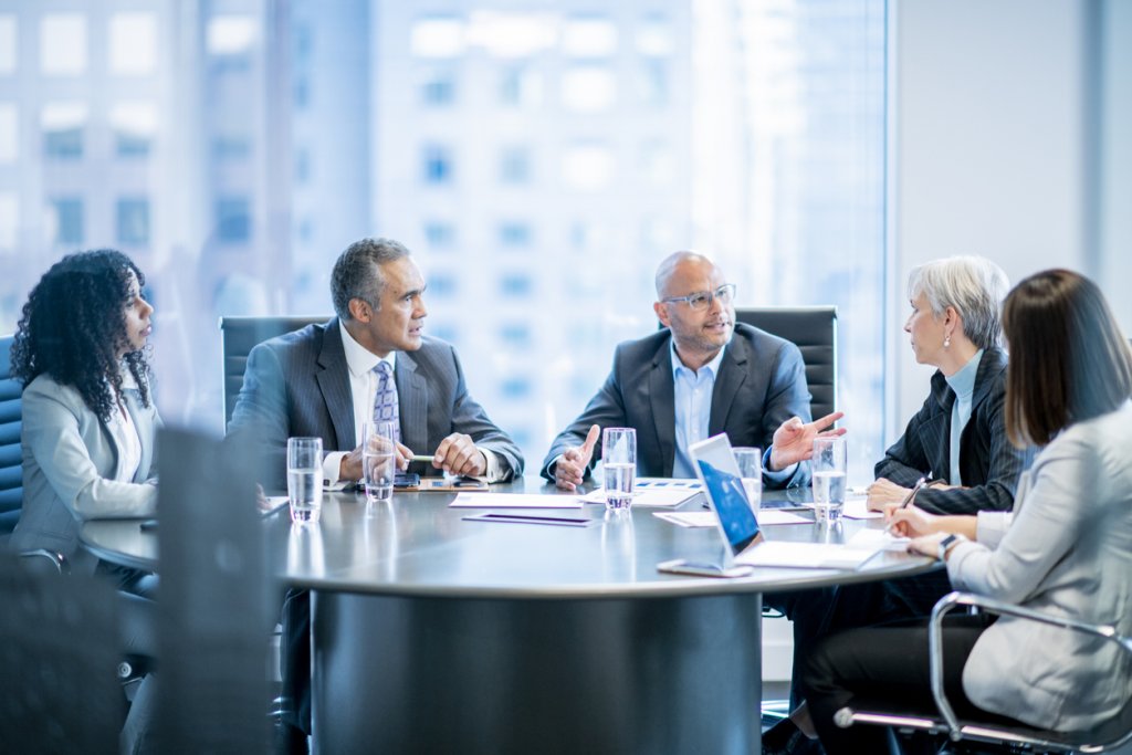 A group of five businesspeople are sitting around an office table. They are talking while having a meeting.